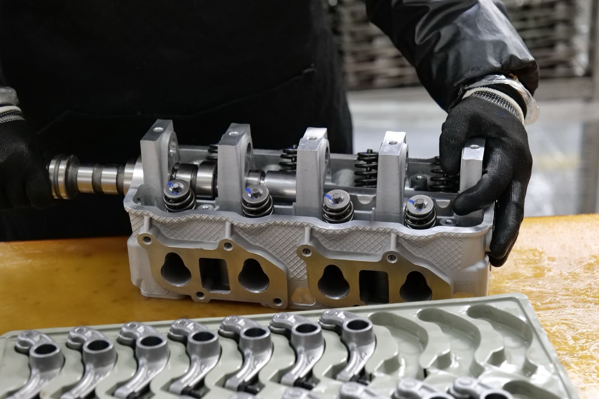 Technician assembling an aluminum cylinder head with camshaft, valve springs and a tray of rocker arms