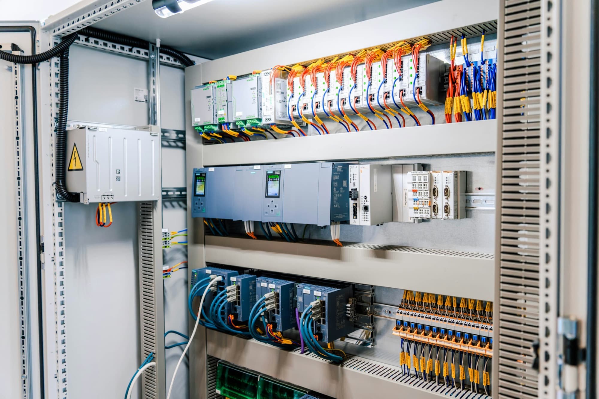 Open industrial control cabinet with PLC, power supplies, network switches and terminal blocks on DIN rail