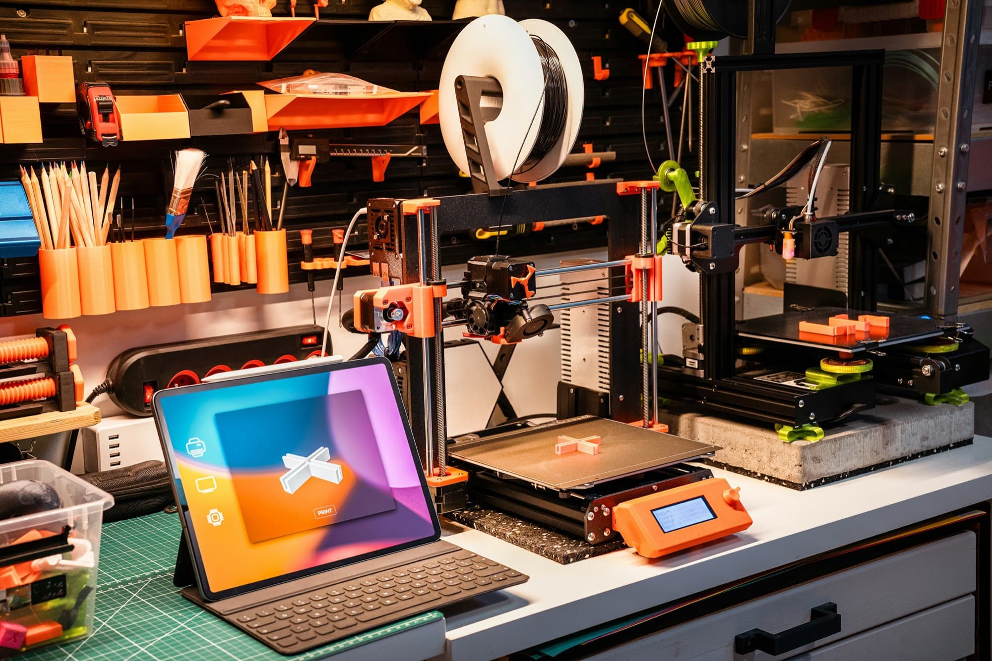 Prototyping workbench with two desktop 3D printers, printed test parts, filament spool and tablet