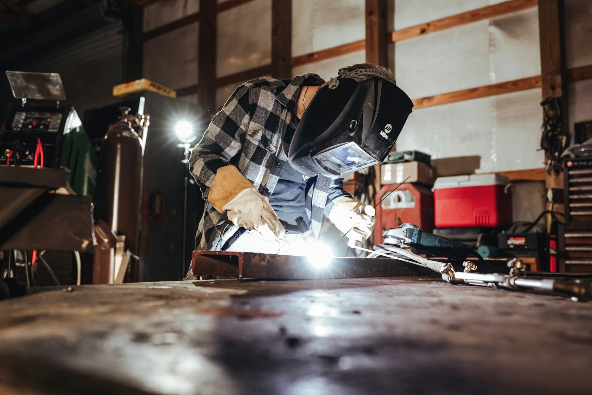 Welder in helmet and gloves TIG welding a steel channel on a workbench in a fabrication shop