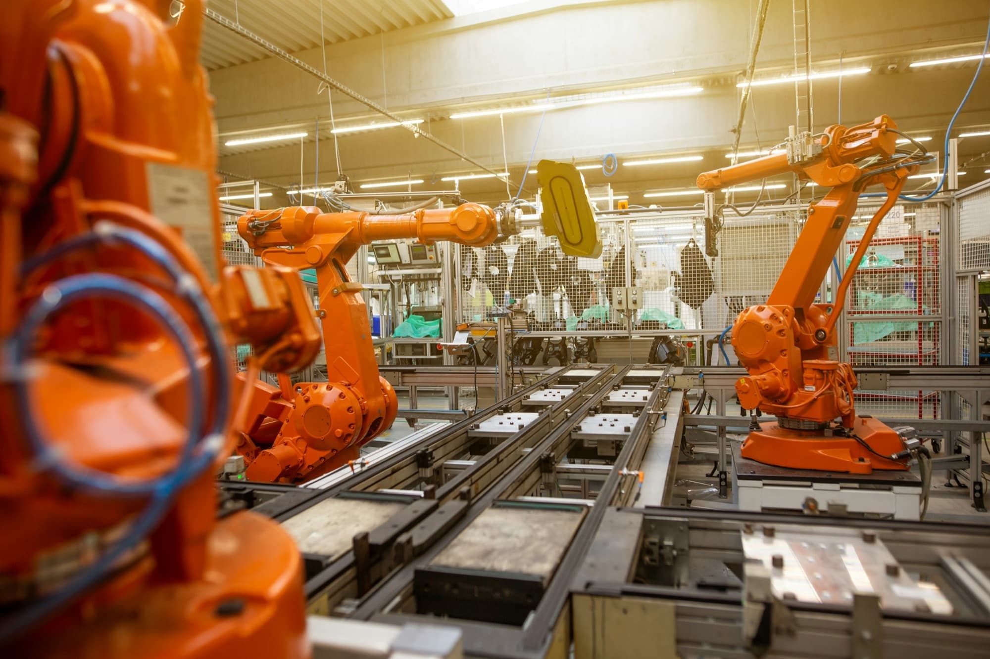 Orange six-axis industrial robots handling parts beside a pallet conveyor inside a fenced work cell