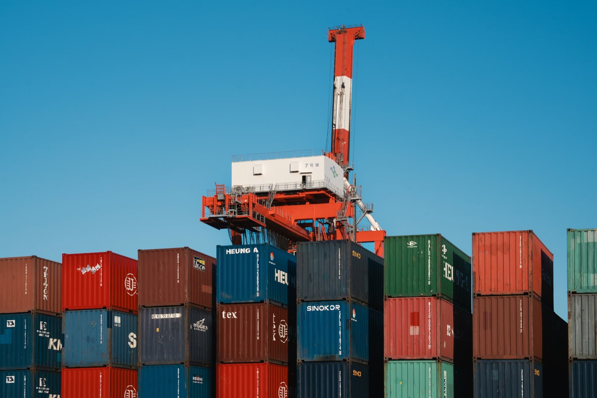 Stacks of shipping containers under a harbor crane against a clear blue sky at a container terminal
