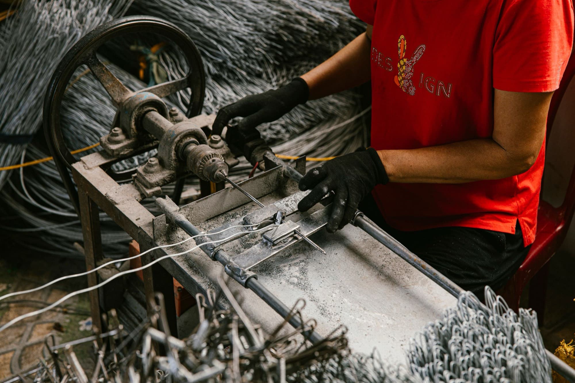 Gloved operator feeding steel wire through a forming jig, with finished wire forms piled in front