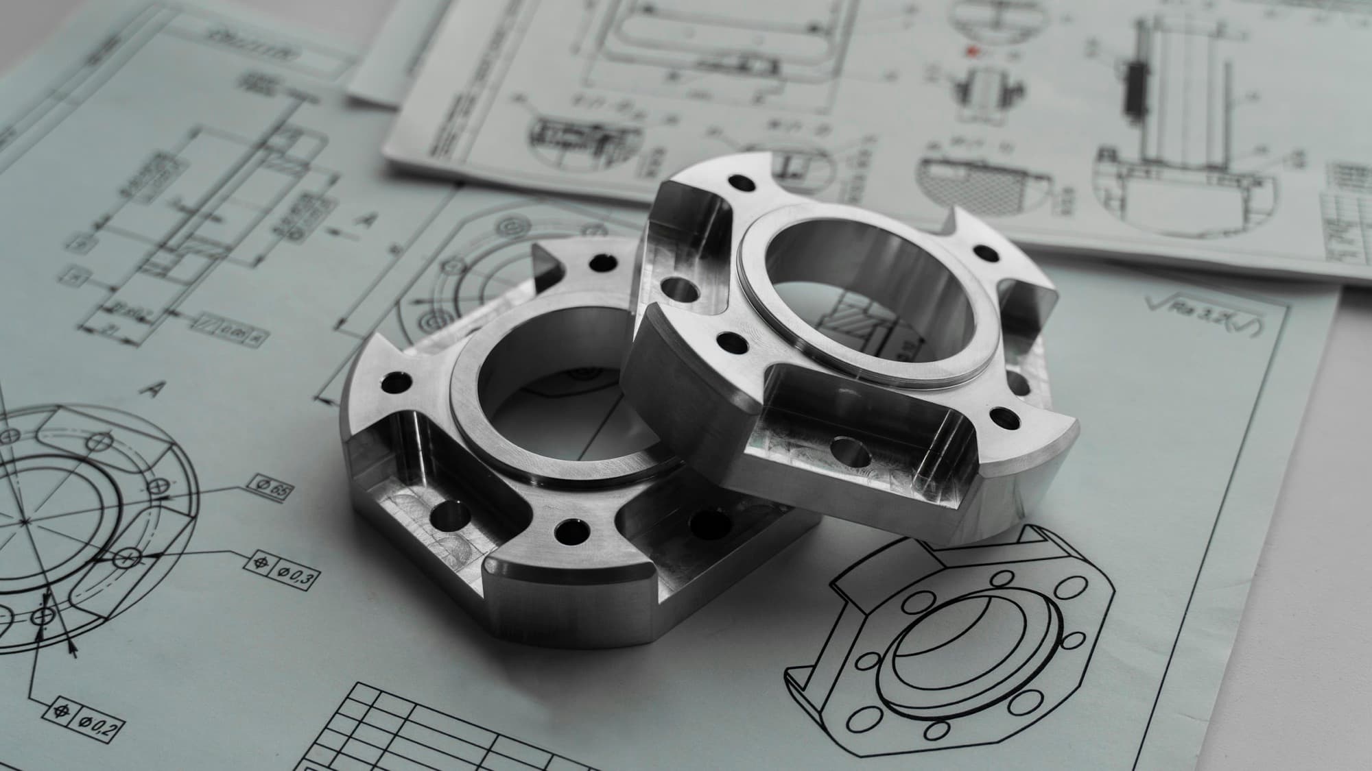 Two machined metal flanges resting on engineering drawings with dimensions and GD&T callouts