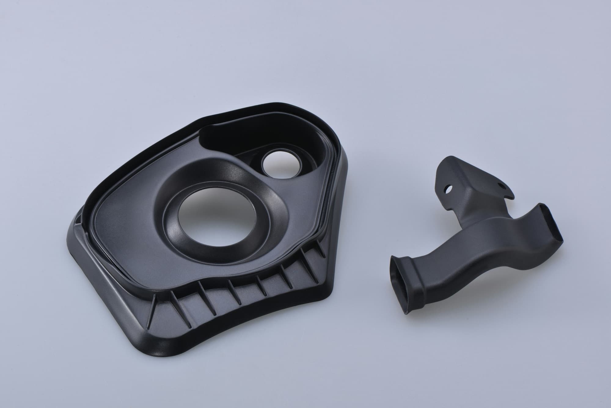Two black injection-molded plastic parts with ribs, bosses and a molded duct on a grey sweep