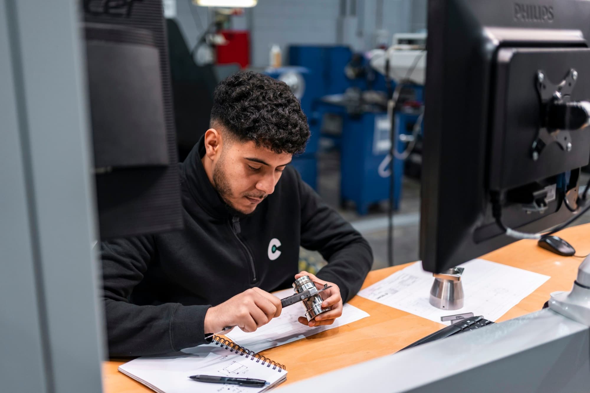 Technician measures a turned stainless part with a caliper against the drawing at an inspection bench