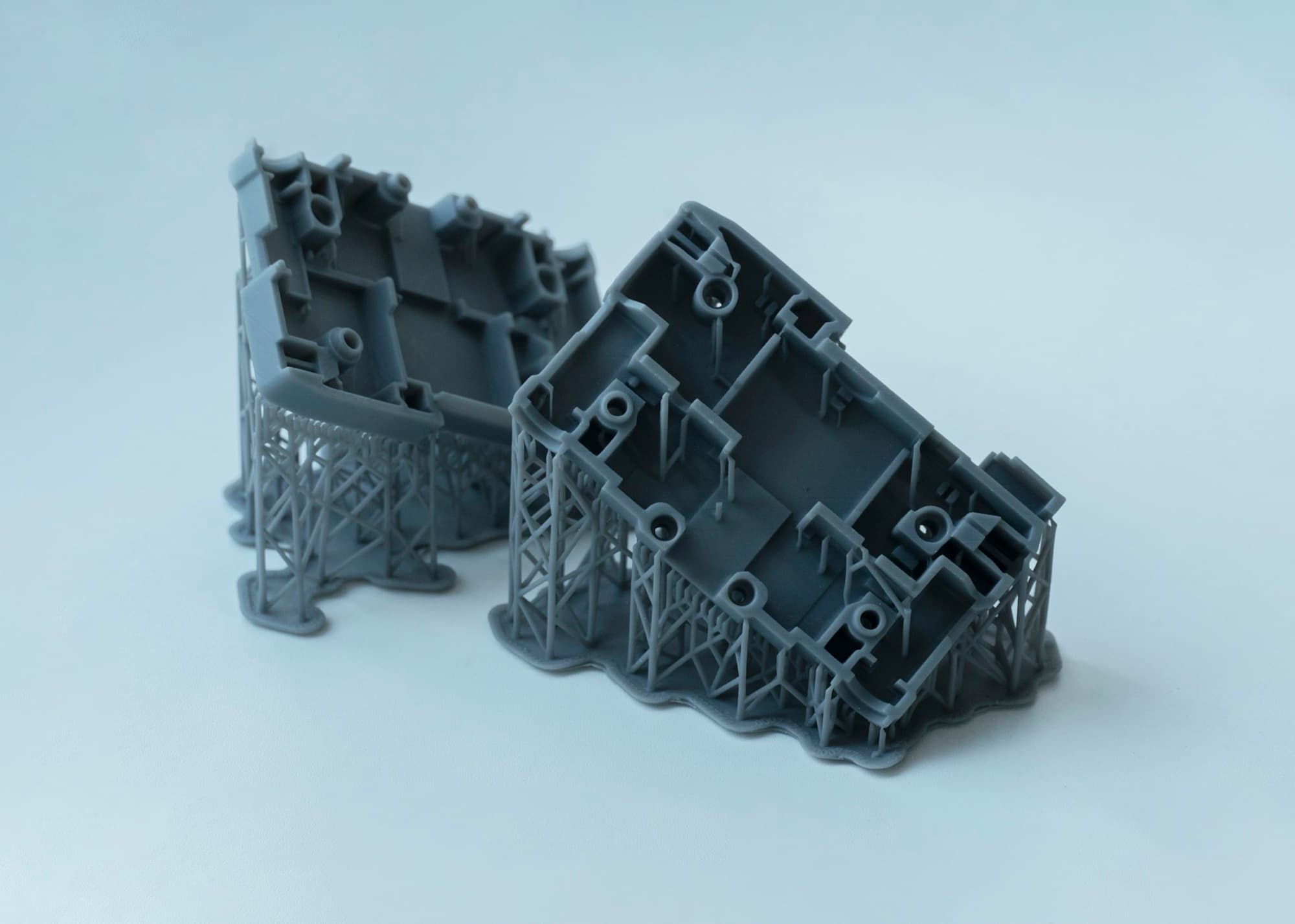 Two resin 3D-printed prototype housings with screw bosses and ribs, still standing on their print supports