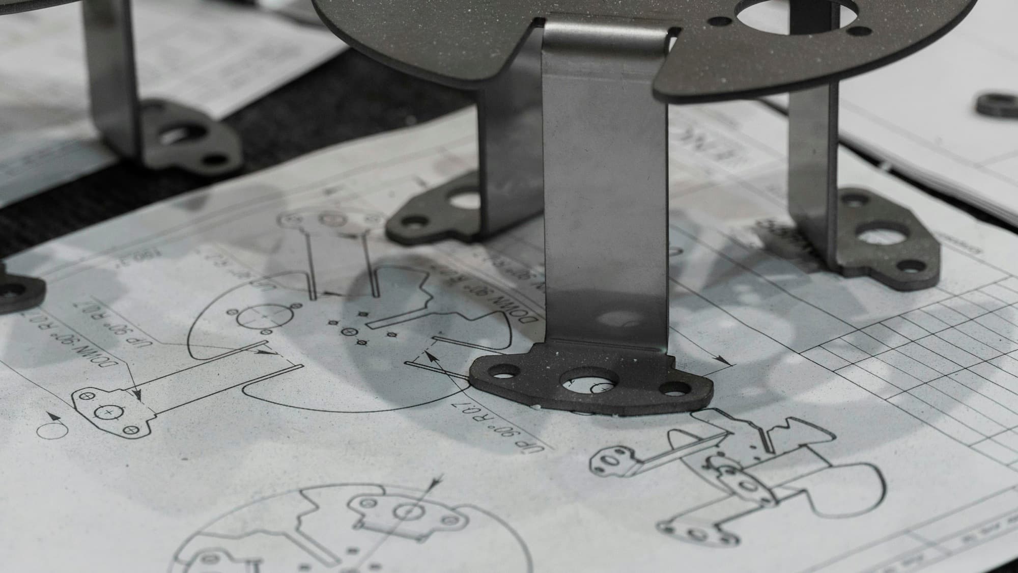 Laser-cut and bent prototype steel brackets standing on the flat-pattern drawing with bend callouts