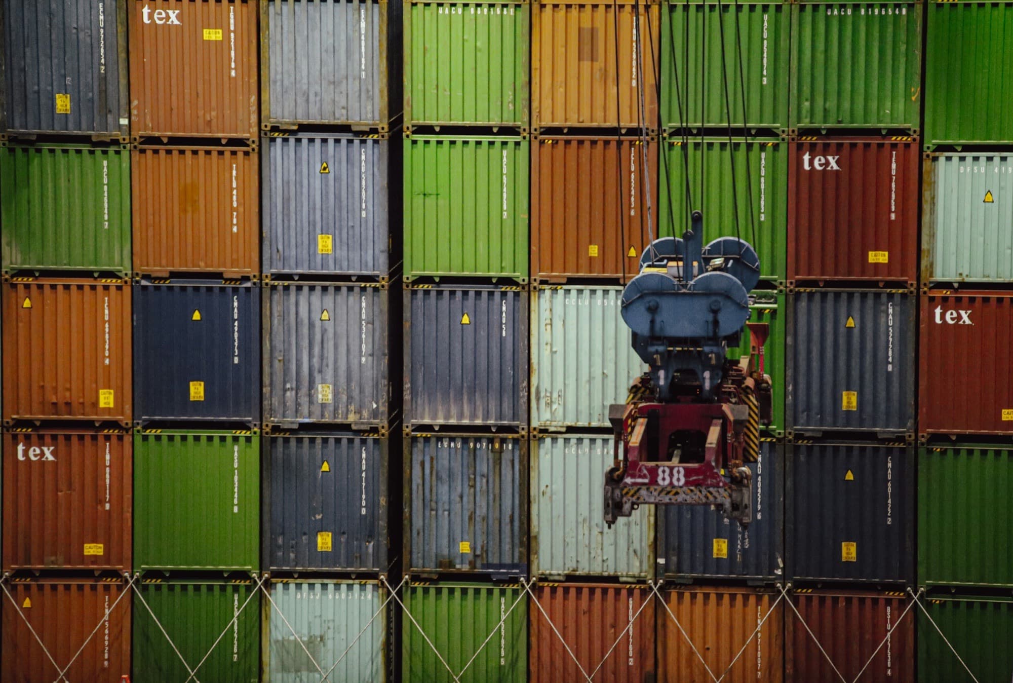 Wall of stacked steel shipping containers with a crane spreader lowering between the stacks