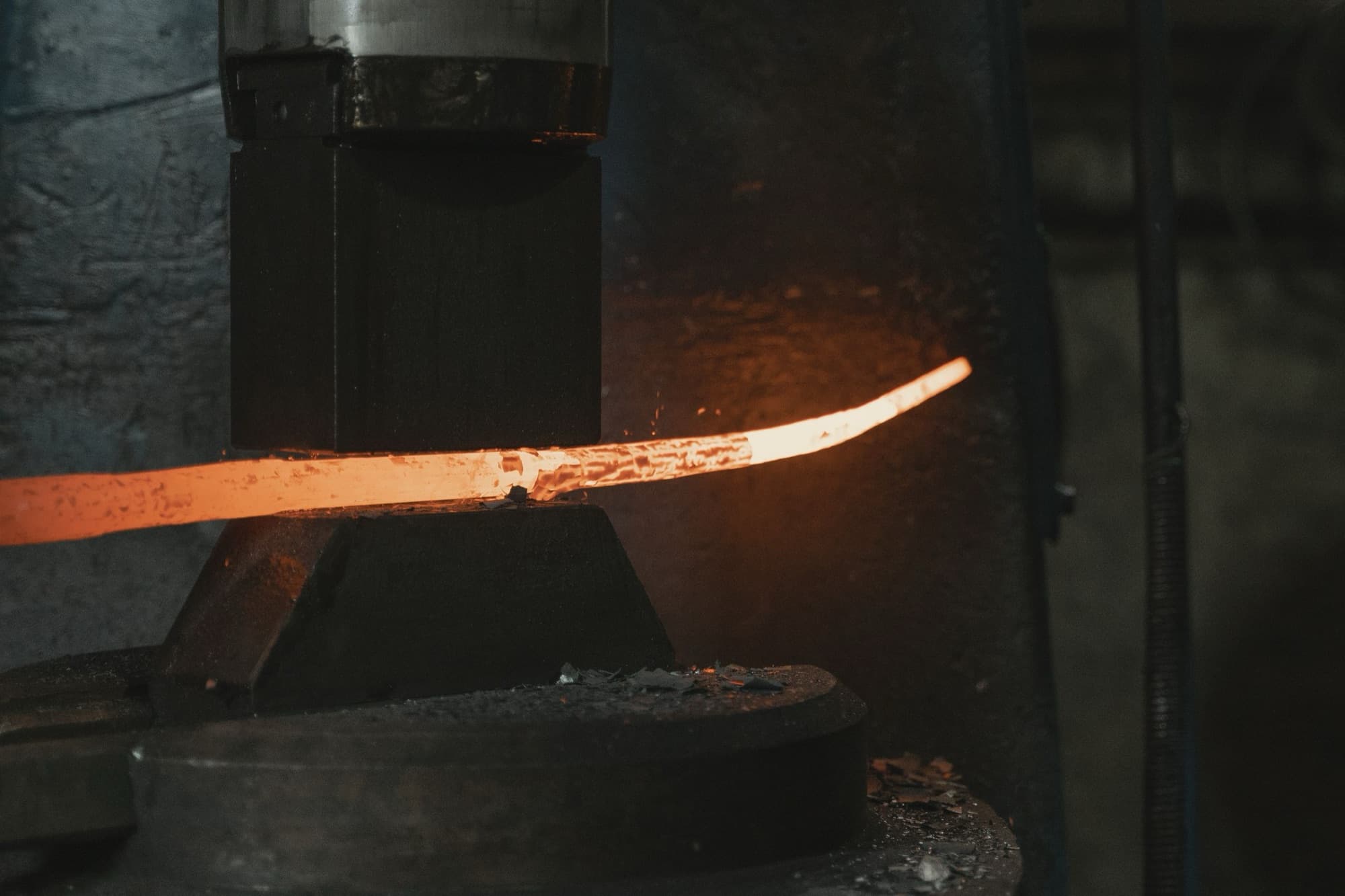 Glowing orange steel bar being shaped between the dies of a forging hammer